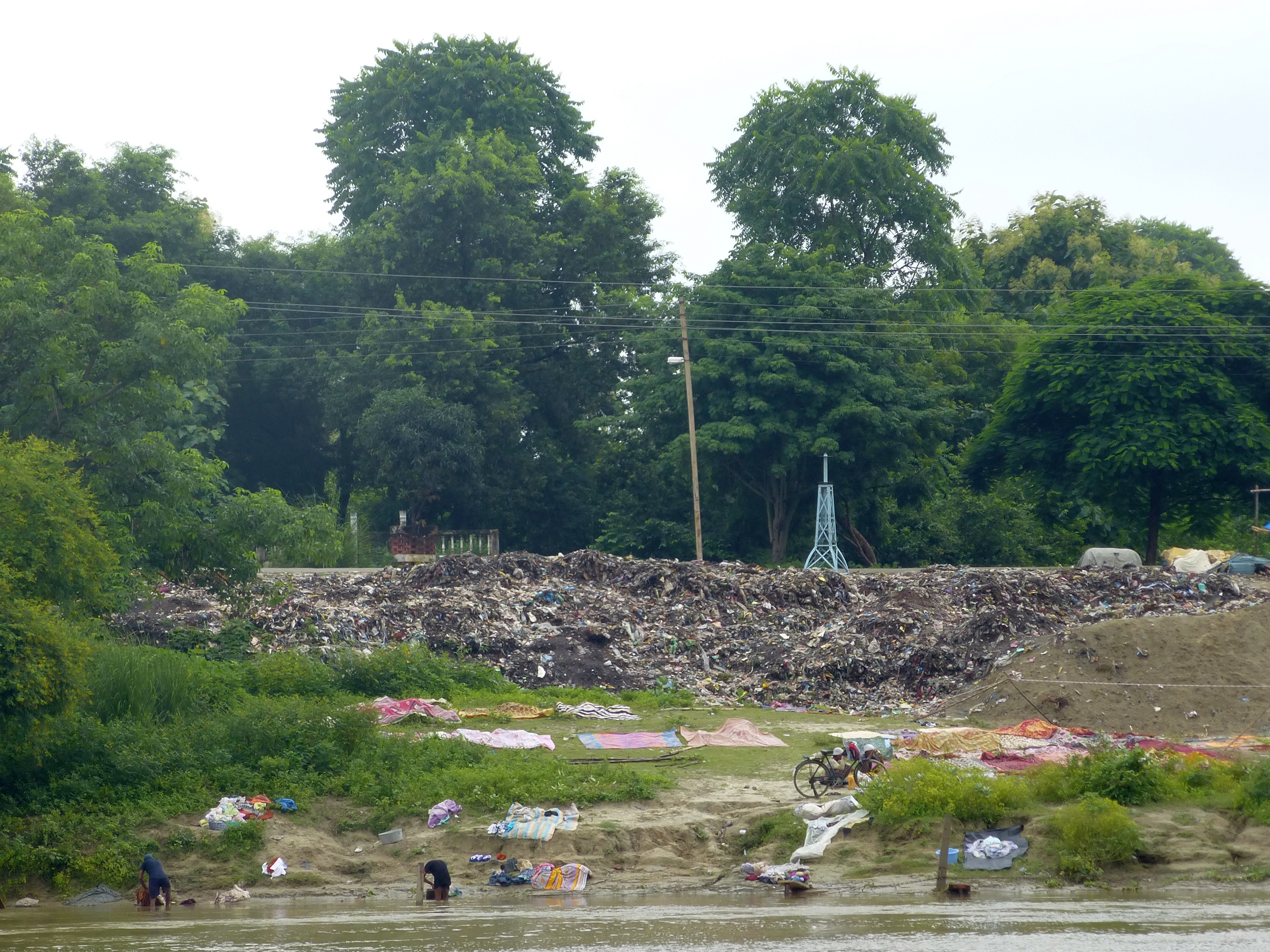 Piles of garbage next to a flowing river where people are washing clothes.
