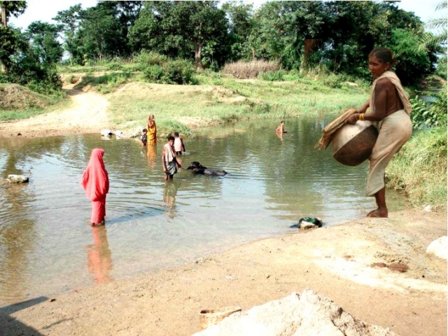 woman collecting drinking water from a pond where others are bathing