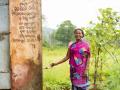 Raibari Bewa standing near the toilet, bathroom unit and collecting water from the third tap in Dudukaguda village, in Thuamul Rampur block, Kalahandi district of Odisha. On the walls, details of Swachh Bharat Mission benefits availed by her in Odia | Picture courtesy: Ajaya Behera